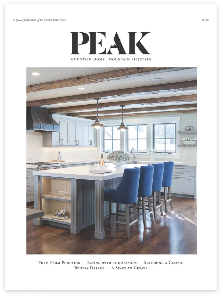 Magazine cover of PEAK featuring a mountain-style kitchen with exposed beams, a farmhouse island, and plush blue barstools, emphasizing seasonal and functional design.