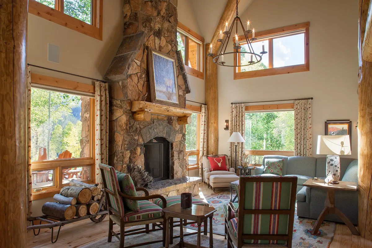 Sunlit living room in mountain home featuring tall stone fireplace and forest views.
