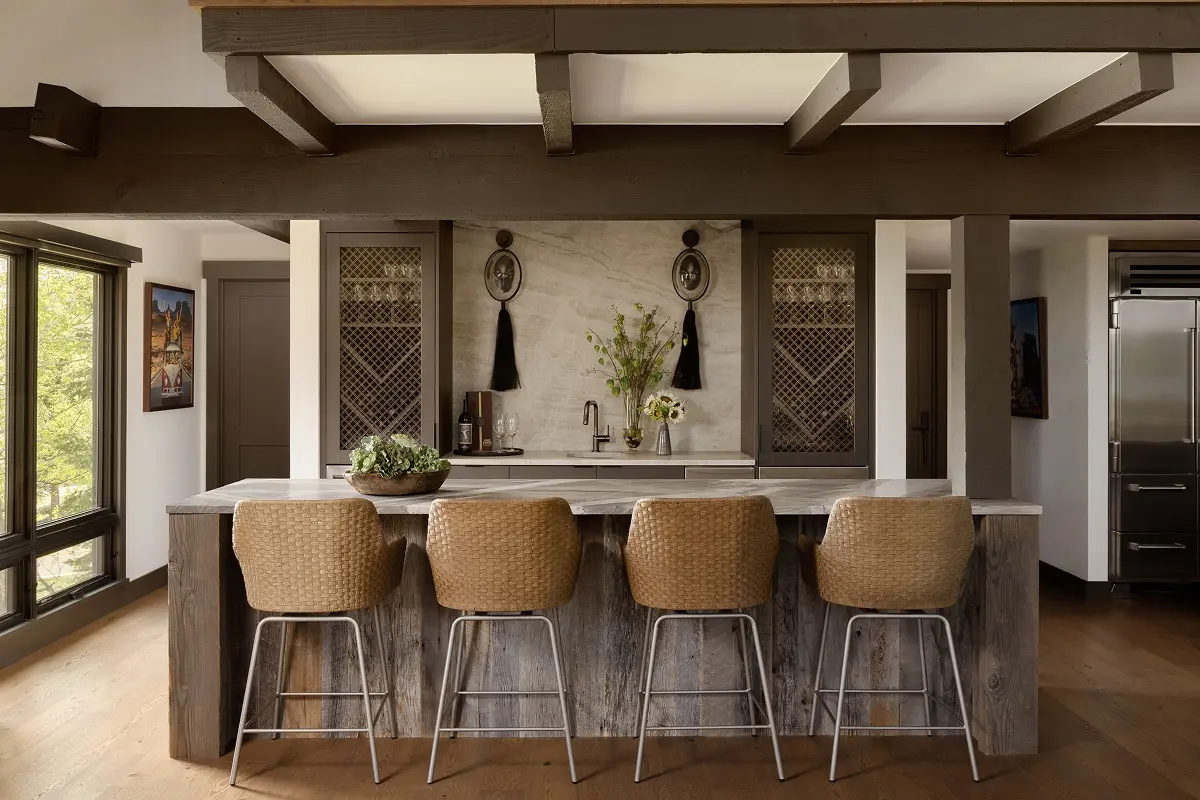 Custom wine bar and prep area with marble backsplash, integrated lighting, and woven barstools in wood-paneled niche.
