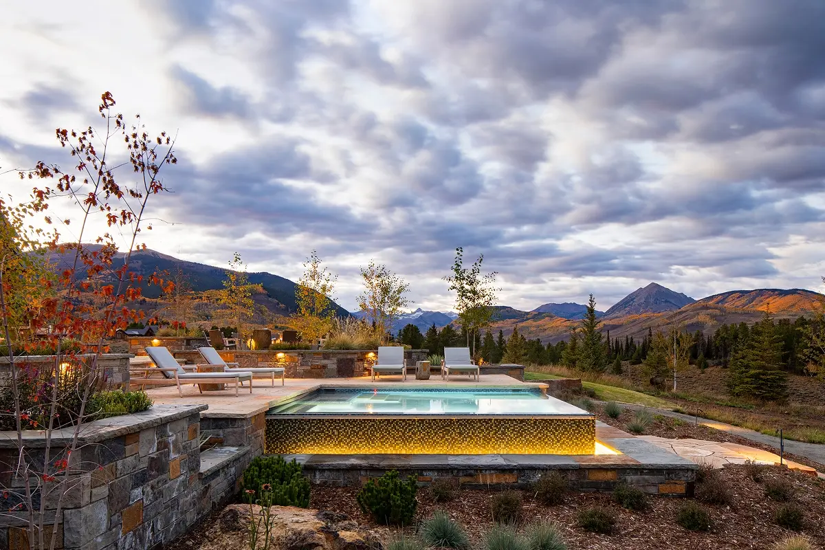 Illuminated infinity-edge pool at dusk, surrounded by lounge chairs and panoramic mountain views.