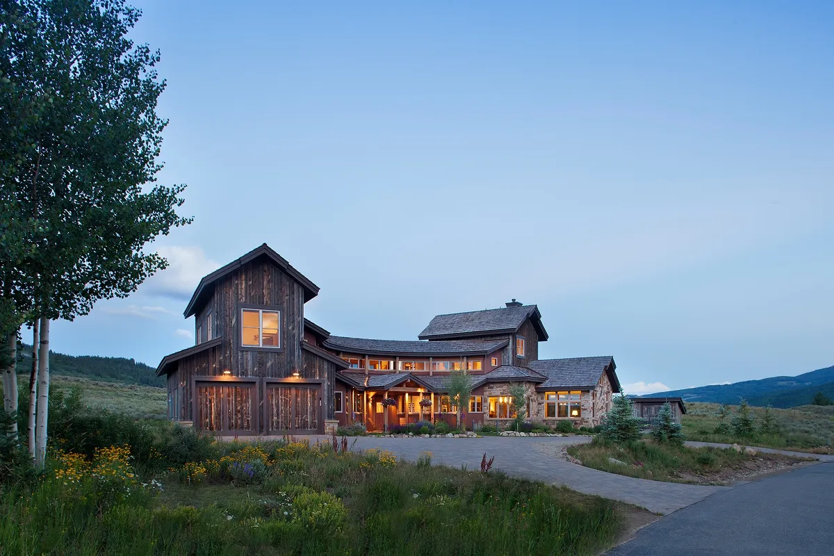 View of mountain home at dusk with lit windows and a stone driveway surrounded by aspen and wildflowers.