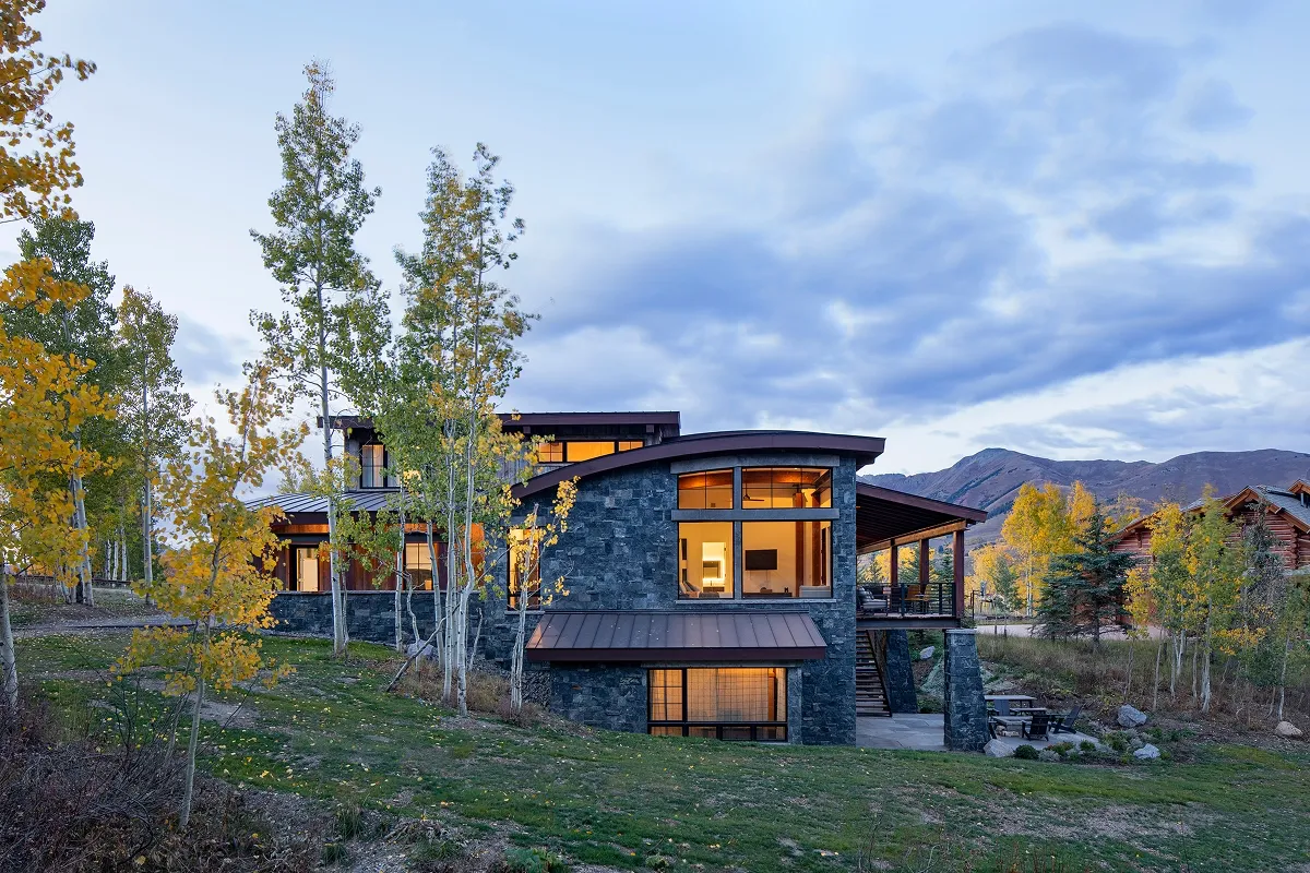 Rear elevation of Surround House at dusk with stone fireplace, large glazing, and sloping roof.