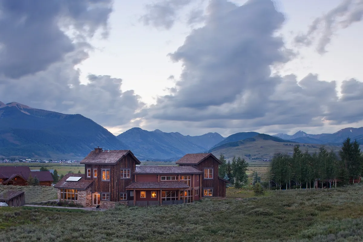 Rear elevation of mountain modern home at dusk with lit windows and surrounding wilderness under dramatic clouds.