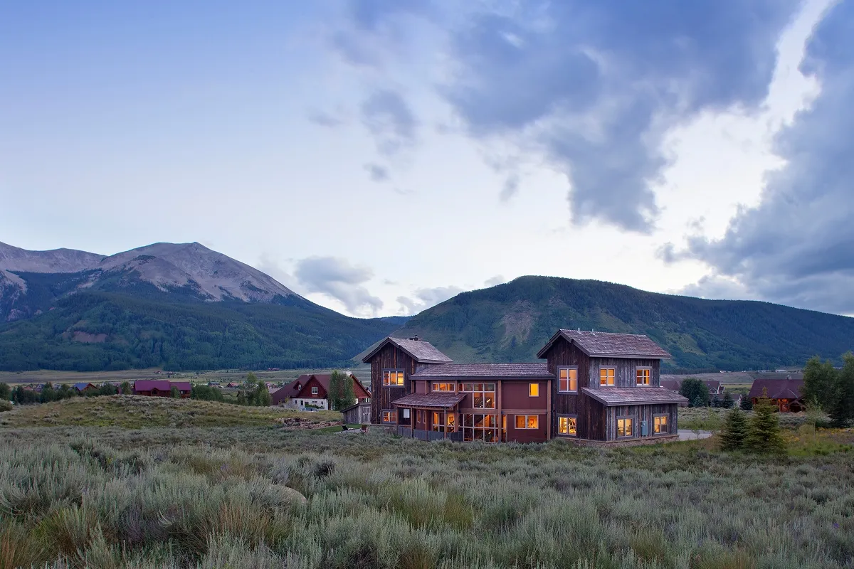 Rear exterior of Crested Butte residence with twilight lighting, mountain views, and rustic wood siding.