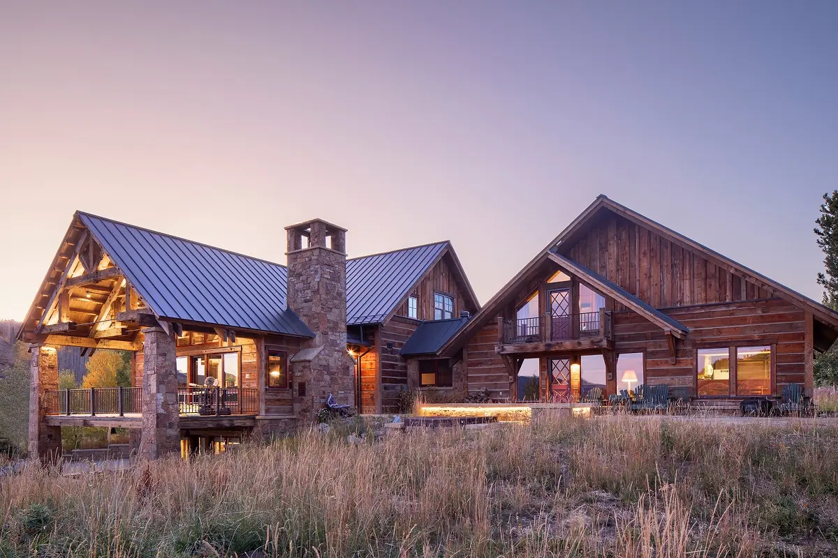 Rustic timber-framed mountain home with stone chimney and warm exterior lighting at dusk.