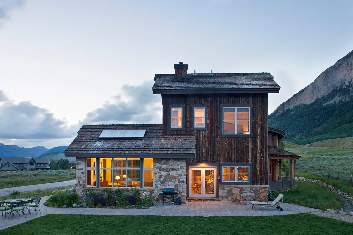 Back view of wood and stone mountain house at dusk with warm interior lighting and outdoor patio space.