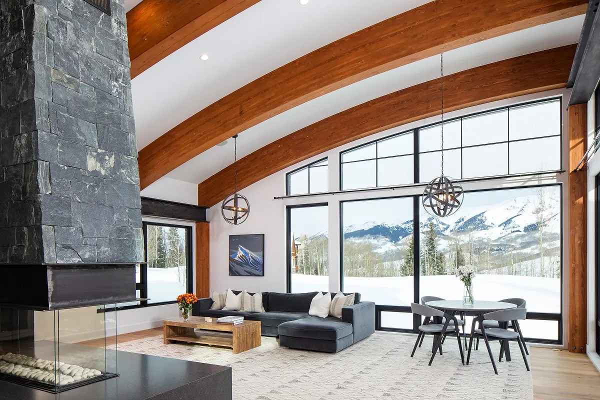Modern mountain living room with arched timber ceiling, stone fireplace, and expansive windows overlooking snowy Crested Butte peaks.